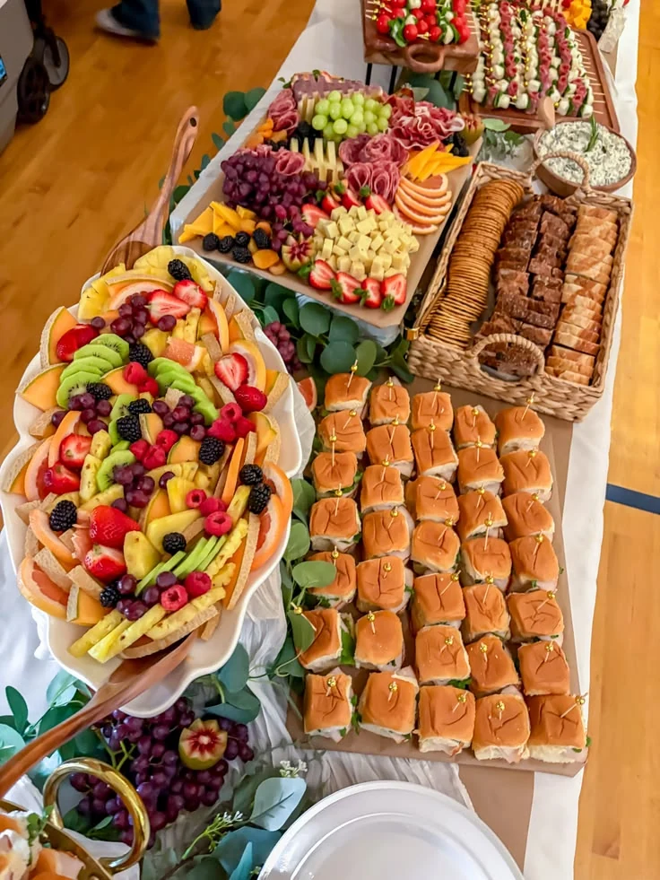 A catered meeting spread with sandwich sliders, fresh fruit, charcuterie, cheeses, crackers, and sweet bites
